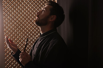 Catholic priest in cassock holding cross and praying to God in confessional
