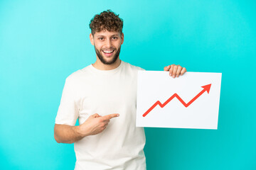 Young handsome caucasian man isolated on blue background holding a sign with a growing statistics arrow symbol with surprised expression