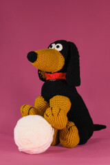 One black and red dachshund crocheted from natural wool and cotton, handmade art. Amigurumi dog doll play with ball on pink background. A soft DIY toy made of cotton.