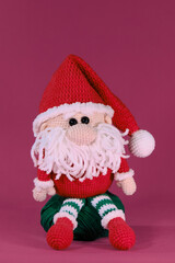 Amigurumi one Christmas gnome with red hat and white beard sits on pink background. Soft DIY toy made of natural cotton and wool. Crocheted santa claus, handmade art. Santas helper.