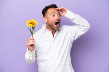 Middle age caucasian man holding sun flower isolated on purple background doing surprise gesture while looking to the side