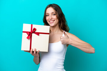 Young caucasian woman holding a gift isolated on blue background with thumbs up because something good has happened