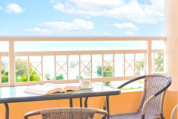 A cup of coffee and an open book over a balcony table. Background view of blue sky and sea. Summer vacation, time for relaxation, hobbies and recreation.