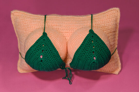 Crocheted pillow in shape of womans breast wears green swimsuit on pink background, handmade art. Female tits without head. Creative gift idea for man.