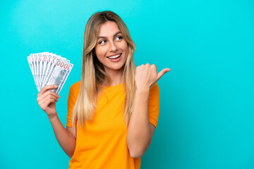 Young Uruguayan woman taking a lot of money isolated on blue background pointing to the side to present a product