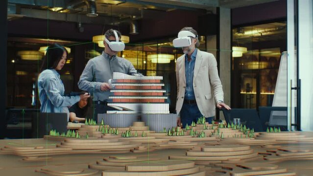 Male architect uses VR headset and wireless controllers, presents modern hi-tech building and park architectural project in virtual reality to colleagues. 3D hologram. Future digital AI technologies.