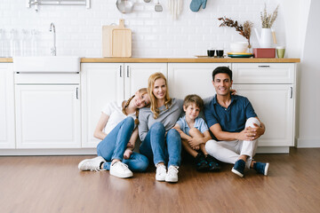 Portrait of happy family father mother daughter son sitting on wooden floor at modern kitchen and looking at camera, spending weekend in cozy apartment, resting at home. Family enjoying weekend time