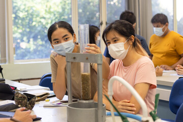 young students with face mask do hydraulic test of samples, note, analyze data using notepad, computer, tablet, phone, calculator in classroom or science laboratory in university