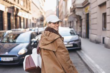 Fototapeta na wymiar Happy girl with blonde hair in light cap and brown trench coat with handbag smiling outdoors, turn around and smiling. Stylish girl in fashion outfit walking in city, girl look back over her shoulder.