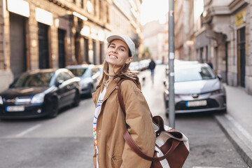 Happy girl with blonde hair in light cap and brown trench coat with handbag smiling outdoors, turn around and smiling. Stylish girl in fashion outfit walking over the city, look happy, hold bag.