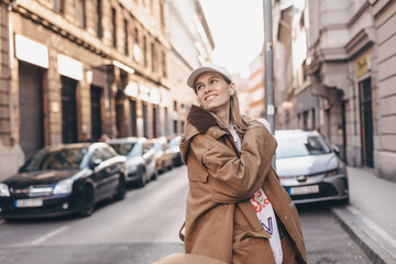 Fototapeta premium Happy girl with blonde hair in light cap and brown trench coat with handbag smiling outdoors, turn around and smiling. Stylish girl in fashion outfit walking over the city, look happy, hold bag.