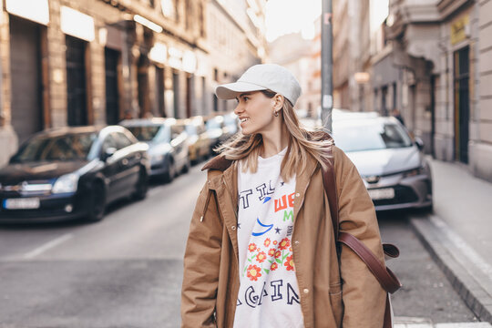 Happy girl with blonde hair in light cap and brown trench coat with handbag smiling outdoors, turn around and smiling. Stylish girl in fashion outfit walking over the city, look happy, hold bag.