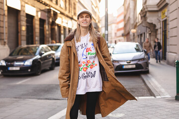 Happy girl with blonde hair in light cap and brown trench coat with handbag smiling outdoors, turn around and smiling. Stylish girl in fashion outfit walking over the city, look happy, hold bag.