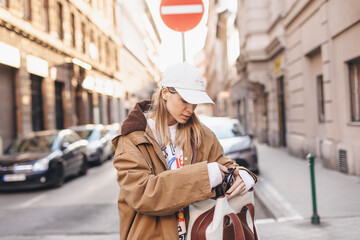 Elegant young woman looking in her brown and white bag her phone. Traveler style woman wear brown trench coat, white cap, sweatshirt and bag on the street. Street style, fashion outfit.