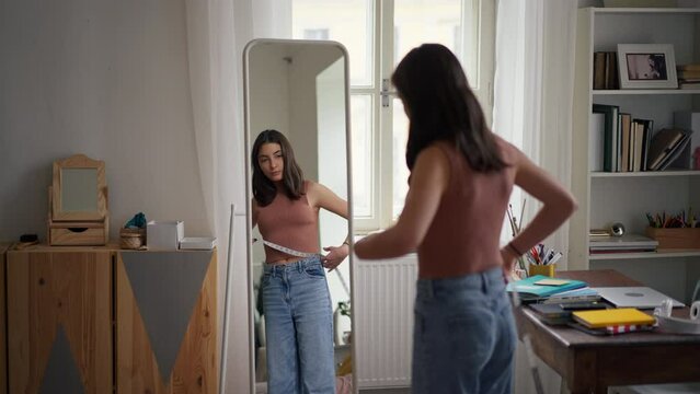 Young teenage girl looking in the mirror in her room, trying on clothes and measuring her waist.