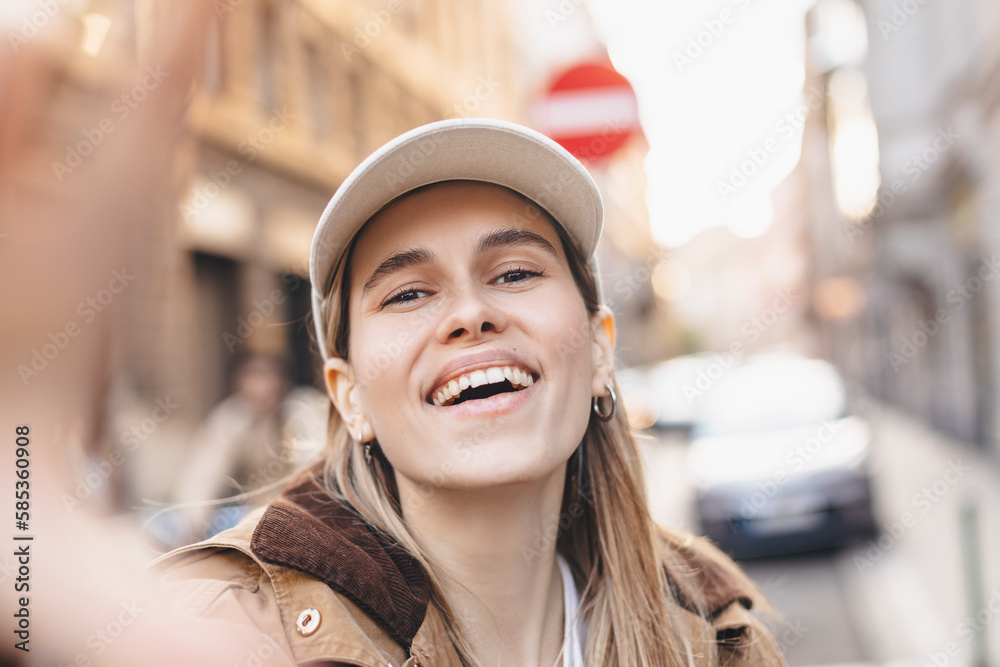 Wall mural Happy young model makes photo holding camera in hand close-up. Woman wear brown trench coat and white cap, look happy, smiling. Outdoor selfie photo in fashion outfit while walking on the street.