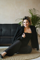 A smiling woman in a black suit is sitting on a rug near a sofa.