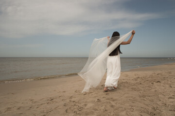girl in white dress with veil walking like a bride on the beach - 585369914