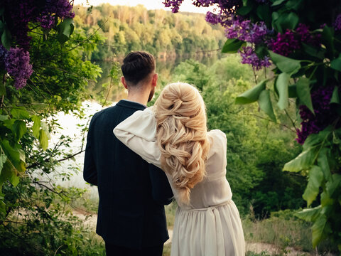 The bride and groom in vintage looks stand at the wedding arch, decorated with lilacs, on the river bank at sunset. Image for your creative design or illustrations.