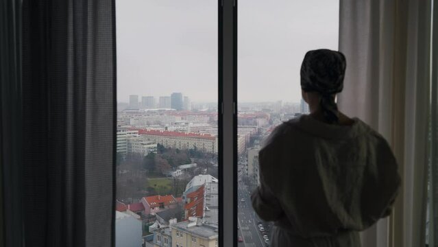Young unhappy woman with cancer standing near a window and looking at view.