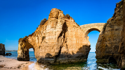 Historic Ponte Romana de Lagos stands gracefully amidst the golden hour glow at Praia dos Estudantes beach, connecting two cliffs and framing the horizon of the Atlantic Ocean in Algarve region.