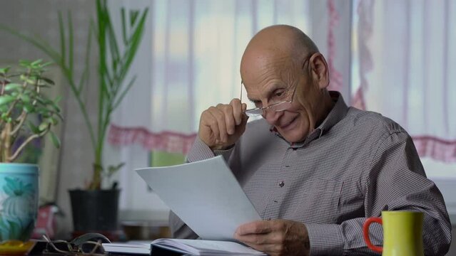 Elderly bald man reads printed article and smiles happily. Senior man enjoys ending work. Old man takes off glasses and put on again at home