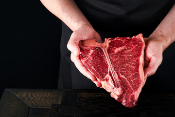 Chef cutting steak beef. Mans hands hold raw steak T-Bone on rustic wooden cutting board on black background. Cooking, recipes and eating concept. Selective focus.