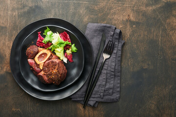 Steaks Osso Buco. Sliced beef grilled Osso Buco or porterhouse meat steak with garnished with salad and french fries on black marble board on old wooden background. Top view. Mock up.