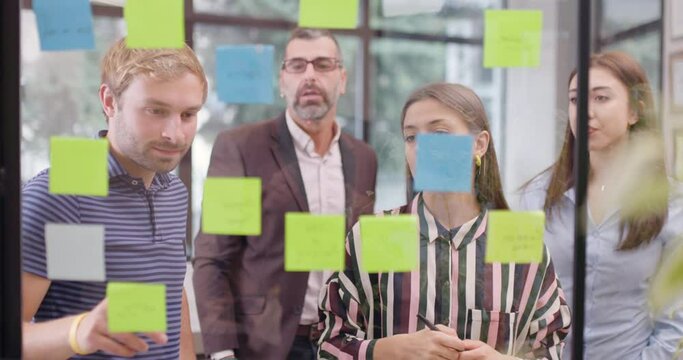 Work colleagues discussing startup project ideas. Team of caucasian business people standing near glass window with sticky notes. Scrum methodology with colorful stickers.