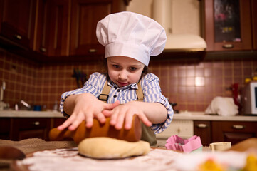 Caucasian lovely little toddler girl in white chef's hat, rolls out dough on floured wooden board, using rolling pin, preparing delicious gingerbread cookies for Easter holidays in the home kitchen