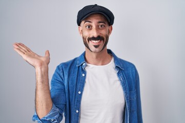 Hispanic man with beard standing over isolated background smiling cheerful presenting and pointing with palm of hand looking at the camera.