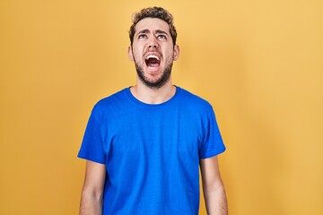 Hispanic man with beard standing over yellow background angry and mad screaming frustrated and...