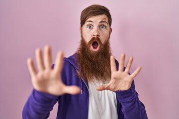 Caucasian man with long beard standing over pink background afraid and terrified with fear expression stop gesture with hands, shouting in shock. panic concept.