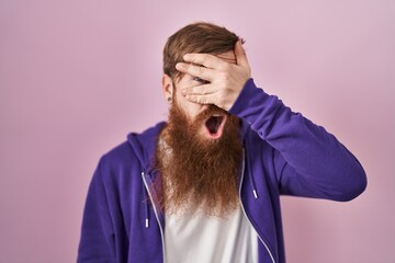 Caucasian man with long beard standing over pink background peeking in shock covering face and eyes with hand, looking through fingers with embarrassed expression.