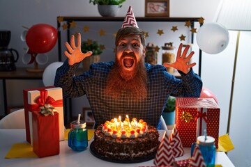 Caucasian man with long beard celebrating birthday holding big chocolate cake celebrating victory with happy smile and winner expression with raised hands