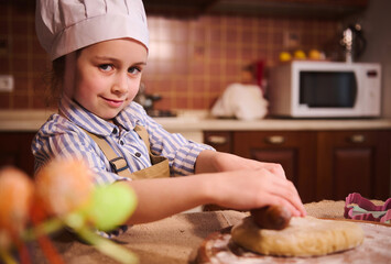 Beautiful little girl 5 years old, wearing white chef's hat and beige apron, smiling cutely looking at camera, standing at kitchen table and rollling out the dough. Blurred Easter eggs on foreground