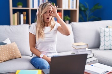 Young blonde woman studying using computer laptop at home doing ok gesture shocked with surprised face, eye looking through fingers. unbelieving expression.