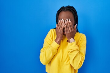 Beautiful black woman standing over blue background rubbing eyes for fatigue and headache, sleepy...