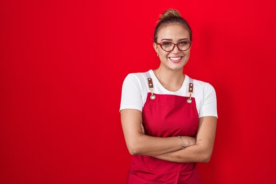 Young hispanic woman wearing waitress apron over red background happy face smiling with crossed arms looking at the camera. positive person.