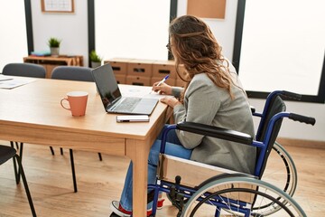 Young beautiful hispanic woman business worker writing on document sitting on wheelchair at office