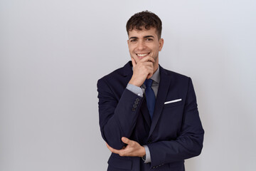 Young hispanic business man wearing suit and tie looking confident at the camera smiling with crossed arms and hand raised on chin. thinking positive.