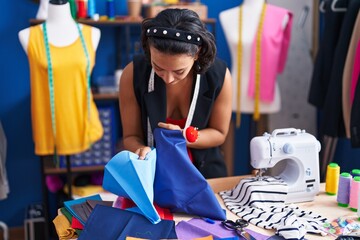 Young beautiful hispanic woman tailor smiling confident holding cloths at clothing factory