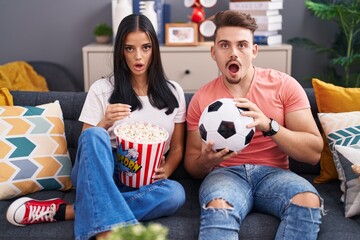 Young hispanic couple football hooligans holding ball and eating popcorn scared and amazed with...
