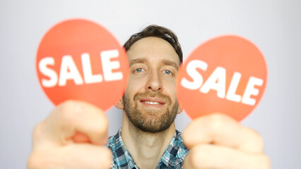Close-up of a cute smiling man showing two defocused red round signs with the word SALE