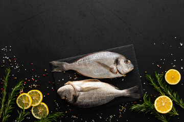 Dorada fish with lemon slices and rosemary sprigs on a slate board among spices on a black background, top view, copy space