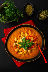 Healthy lunches, tomato and carrot puree soup, beans with pumpkin seeds, healthy tasty legumes, top view of a plate of soup on a black background