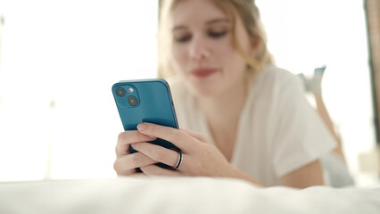 Young blonde woman using smartphone lying on bed at bedroom