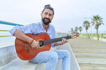 Young hispanic man musician playing classical guitar sitting on bench at seaside