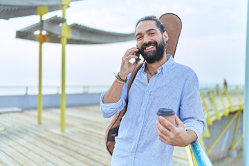 Young hispanic man musician talking on smartphone drinking coffee at seaside