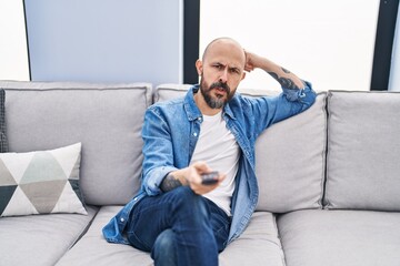 Young bald man watching tv sitting on sofa with boring expression at home